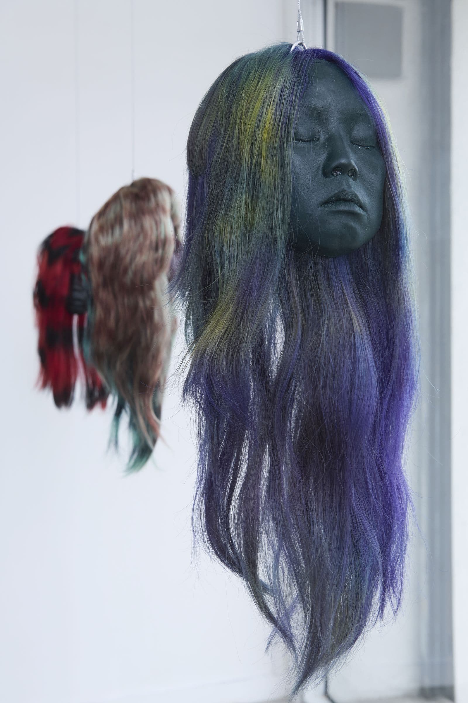 Suspended sculpture with long violet hair in front of a white gallery wall